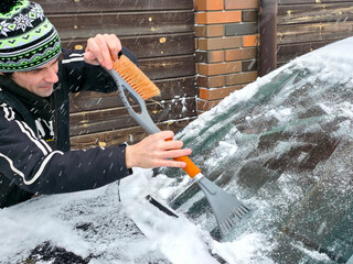Man scraping snow from car windshield with an ice scraper and brush. Winter concept for cold weather, vehicle maintenance, and daily commute preparation.