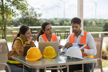 Team of engineers collaborating with digital devices at outdoor office table, planning renewable energy project with wind turbines in background, showcasing teamwork and green technology.