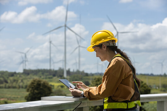 Female engineer wearing safety helmet and vest holding tablet, observing wind turbines on bright day, representing clean energy, innovation, sustainable engineering. - Powered by Adobe