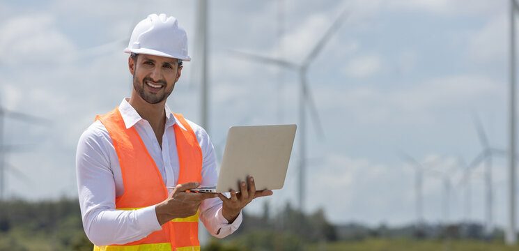 Smiling male engineer in safety vest and helmet using laptop at wind farm, representing renewable energy technology, field operations, sustainable development. - Powered by Adobe