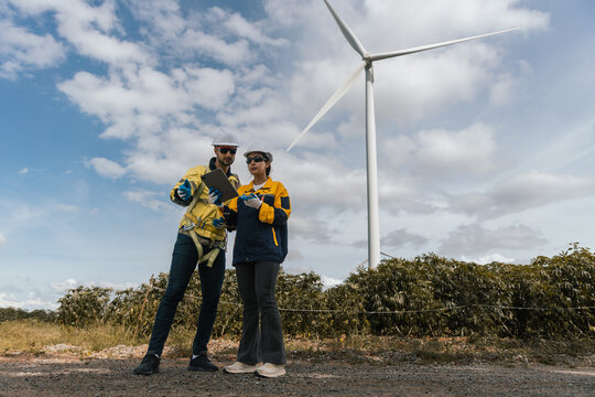 Two wind turbine technicians wearing safety gear analyzing data on tablet in front of wind turbine, symbolizing teamwork, renewable energy, smart field operations. - Powered by Adobe