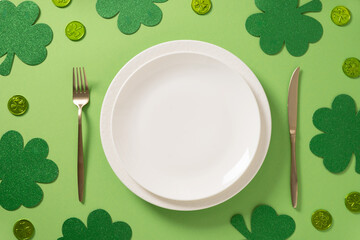 St Patrick's Day table setting with empty white plate and traditional shamrock, clover leaves and coins on light green background. View from above.
