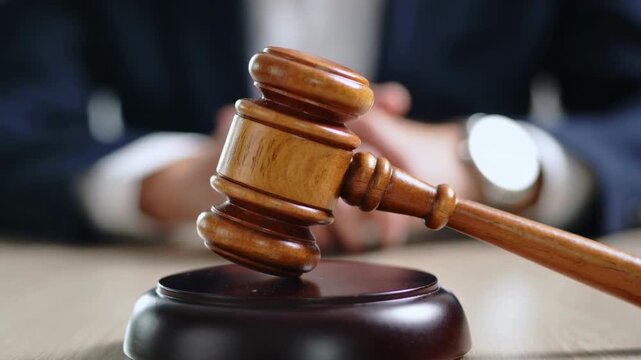 The image shows a wooden gavel on its sound block, with a person in the background. The gavel is in focus, suggesting a legal or judicial context.