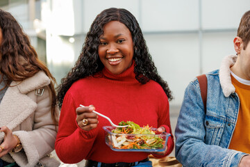 Smiling young student eating healthy salad with diverse friends during a college lunch outdoors