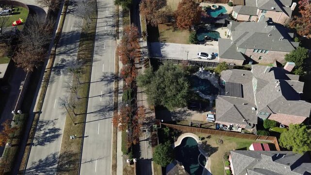 Wide road with bare trees divides two residential sections in Coppell, Texas. Homes feature fenced yards, driveways, pools. Seasonal leaf drop and evergreen contrast mark fall-winter transition