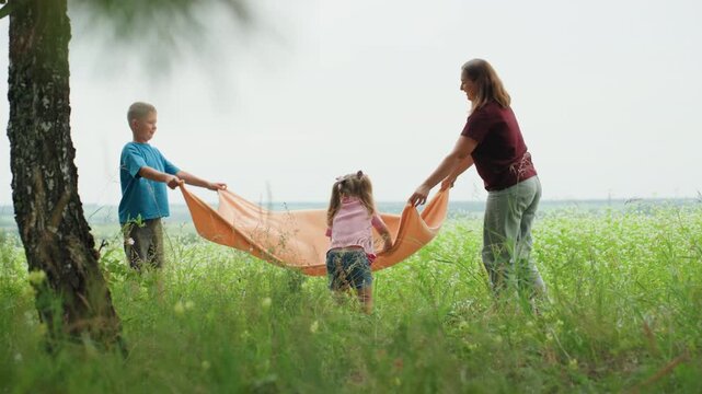 white mother leads playful parachute game, children lift orange blanket into shelter and crawl under then pop out laughing, birch tree and tall grass frame scene, bright summer sunlight and spirited