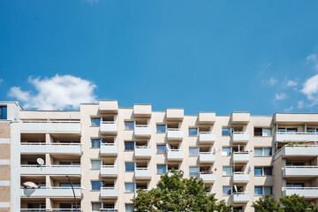 Low angle view of a modern housing complex facade featuring rows of balconies and windows under a bright blue sky with green trees at the base.