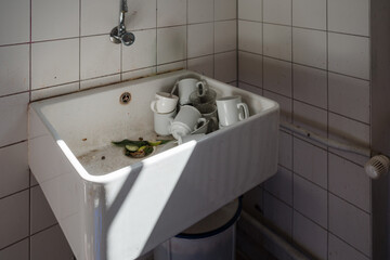 A high angle view of a deep white ceramic utility sink located in a tiled corner. The basin is cluttered with dirty white coffee mugs, drinking glasses, and food waste including cucumber slices and ci