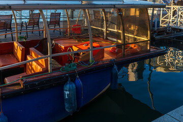 Small covered passenger boat tied to a riverside dock, reflected in calm water during warm evening light. Travel and transportation concept showing leisure boating, urban river life, and quiet harbor 