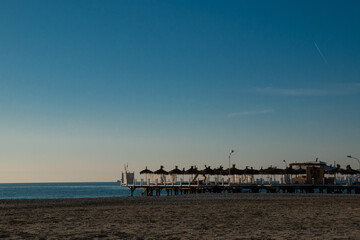 A quiet, empty beach with a wooden pier and umbrellas on the calm sea.