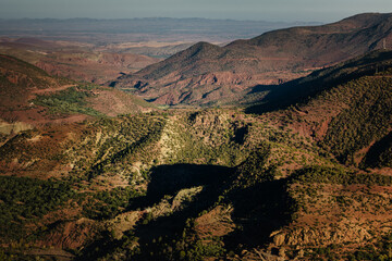 Sunlit mountain valleys in Morocco&rsquo;s Atlas region, with layered ridges, scattered vegetation, and deep shadows revealing the scale and texture of the rugged highland landscape.