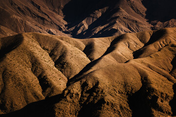 Sunlit ridges and deep shadows shape the arid slopes of the Atlas Mountains in Morocco, revealing erosion patterns and dramatic textures of a rugged, almost abstract landscape.