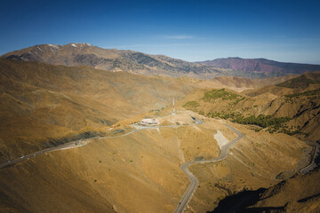 Winding mountain road crossing the High Atlas in Morocco, with arid ridges, scattered greenery, and distant peaks under a clear blue sky, showing scale and isolation of the landscape.