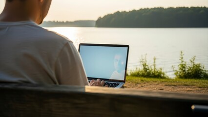 Man working on laptop by a serene lake at sunset, enjoying remote work outdoors.