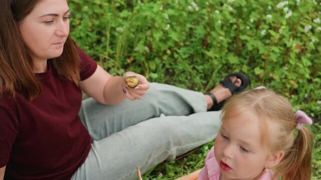 white mother feeding daughter snack on picnic blanket in meadow mother offers breadstick from wicker basket while daughter tastes and looks away playful pigtails pink outfit candid family bond