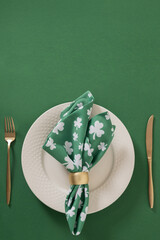 St Patricks day festive table setting with white plate, green shamrock napkin on green background. Vertical format. Template. Flat lay, holiday celebration.