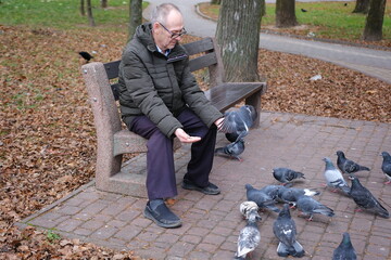 An elderly man enjoys a cold winter morning in the city. A gray-haired man feeds pigeons while sitting on a bench in the square. High quality photo