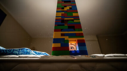 Boy peeking through colorful toy structure on bed, illuminated by soft lighting in a cozy bedroom setting, building