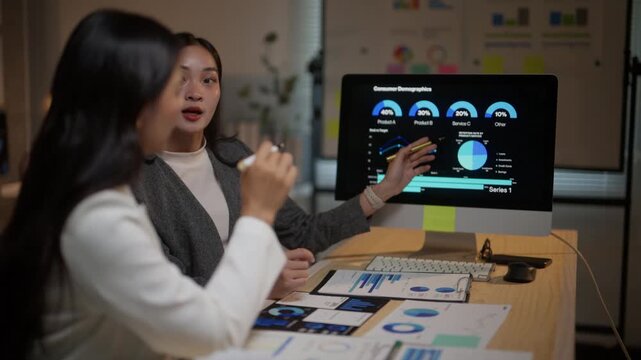 Young asian businesswomen working together in the office late at night. One woman explains consumer demographics data on a computer screen
