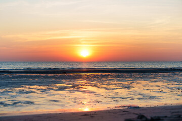 Obraz premium A vivid sunset over the ocean with warm orange and golden tones, gentle waves, and reflections on wet sand, creating a peaceful coastal scene at dusk with a calm horizon.