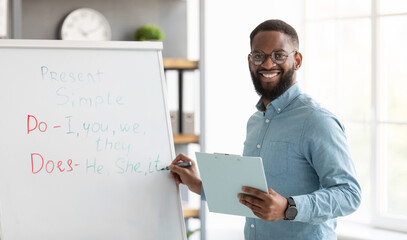 Smiling millennial african american bearded guy teacher in glasses stands near blackboard and...