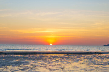 Fototapeta premium Golden sunset over a calm ocean with the sun touching the horizon, warm orange sky reflected on wet sand, creating a peaceful and serene seaside landscape.