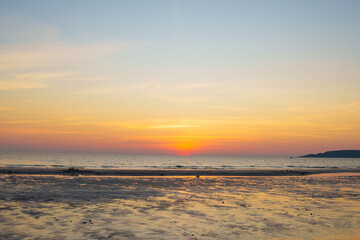 Fototapeta premium Golden sunset over a calm ocean with the sun touching the horizon, warm orange sky reflected on wet sand, creating a peaceful and serene seaside landscape.