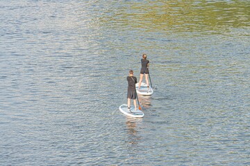 Tranquil scene of two paddleboarders on calm water, casual sportswear, long narrow boards, natural background with trees and foliage, late afternoonearly evening soft glow