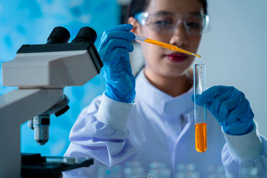 A woman wearing a lab coat and safety glasses is looking through a microscope. She is focused on the image in front of her, likely studying something related to science or research