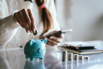 Blue piggy bank with stacked coins on a table, while a person uses a calculator in the background, representing saving money, budgeting, personal finance, and financial planning concepts.