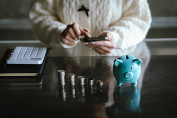 Blue piggy bank with stacked coins on a table, while a person uses a calculator in the background, representing saving money, budgeting, personal finance, and financial planning concepts.