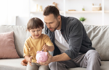 Early financial education. Cute little boy putting coin into pink piggy bank, careful daddy...