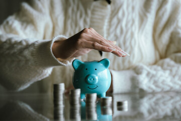 Blue piggy bank with stacked coins on a table, while a person uses a calculator in the background, representing saving money, budgeting, personal finance, and financial planning concepts.