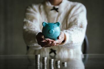 Blue piggy bank with stacked coins on a table, while a person uses a calculator in the background, representing saving money, budgeting, personal finance, and financial planning concepts.