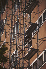 Scaffolding stands beside a building with multiple windows under clear skies. Workers are likely involved in repair or construction tasks on upper levels in the city