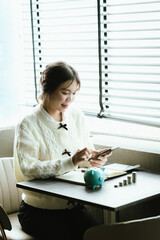 Young Asian woman calculating personal finances at home, using a calculator beside a piggy bank and coins on a desk by the window, symbolizing budgeting, saving money, and financial planning.