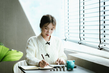 Young Asian woman calculating personal finances at home, using a calculator beside a piggy bank and coins on a desk by the window, symbolizing budgeting, saving money, and financial planning.