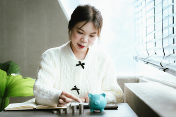 Young Asian woman calculating personal finances at home, using a calculator beside a piggy bank and coins on a desk by the window, symbolizing budgeting, saving money, and financial planning.