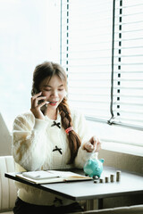 Young Asian woman calculating personal finances at home, using a calculator beside a piggy bank and coins on a desk by the window, symbolizing budgeting, saving money, and financial planning.