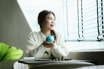 Young Asian woman calculating personal finances at home, using a calculator beside a piggy bank and coins on a desk by the window, symbolizing budgeting, saving money, and financial planning.
