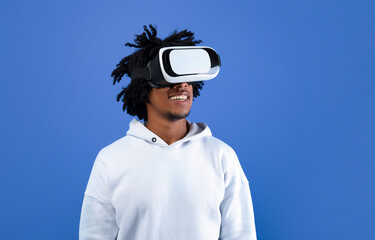 Cool African American guy wearing VR headset, using virtual reality for entertainment on blue studio background, copy space. Funky black man exploring cyberspace. Futuristic technologies