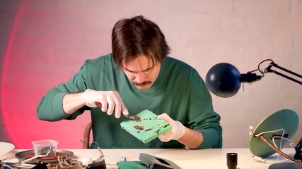 Man restoration expert repairing electric item at table, disassembling vintage lamp, holding base and scraping off paint with putty knife. Craftsman in gloves taking off layers of paint from lamp base