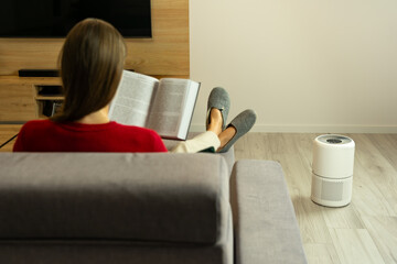 rear view of a woman reading a book while sitting on a gray sofa, with air purifier in a minimalist interior. Enjoy cleaner, healthier air at home. Four-layer HEPA filtration system