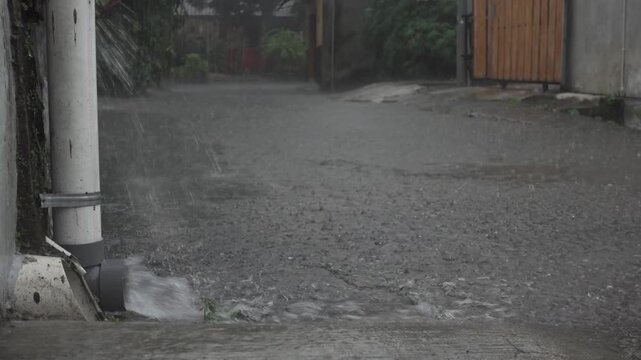 Heavy rainwater pouring from a roof gutter pipe in front of a house, capturing intense rainfall and water drainage during a stormy weather condition.