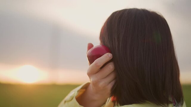 Serene woman holding apple during sunset, Contemplative woman with apple gazing into fading sunset sky, Peaceful female figure cradling apple as evening light bathes rural landscape in tranquility