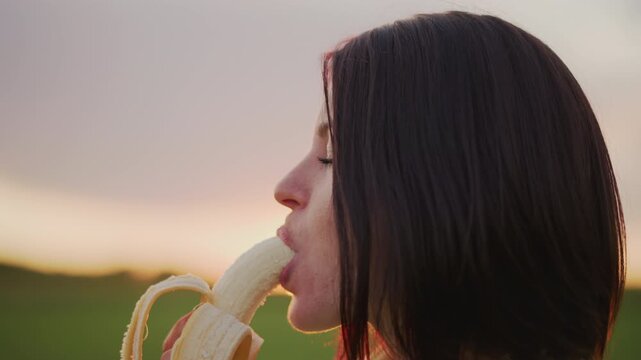 Caucasian woman eating banana in meadow profile shot with sunset rim light, slow living mood, side view portrait, braid and soft hair silhouette, quiet savoring and mindful chewing, pastoral horizon