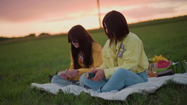 sunset picnic two women on blanket in meadow, sharing snacks and playing small handheld instruments, soft twilight glow, cinematic warm tones, relaxed intimate conversation, basket with fruit