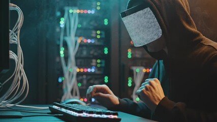 Hooded hacker types on keyboard in server room with datacenter lights. Cybercriminal works typing in dark server datacenter. Hacker in hood uses keyboard near server cables and network lights. - Powered by Adobe