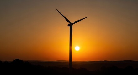 Obraz premium A wind turbine silhouetted against a vibrant orange and yellow sunset sky, with a mountain range in the background.