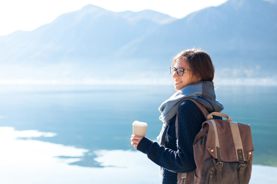 Happy woman traveling by mountain lake, drinking coffee. Traveler enjoying winter sea beach, sabbatical trip for mental health, adventure, relaxing. Girl with backpack on coast. Solo female tourism